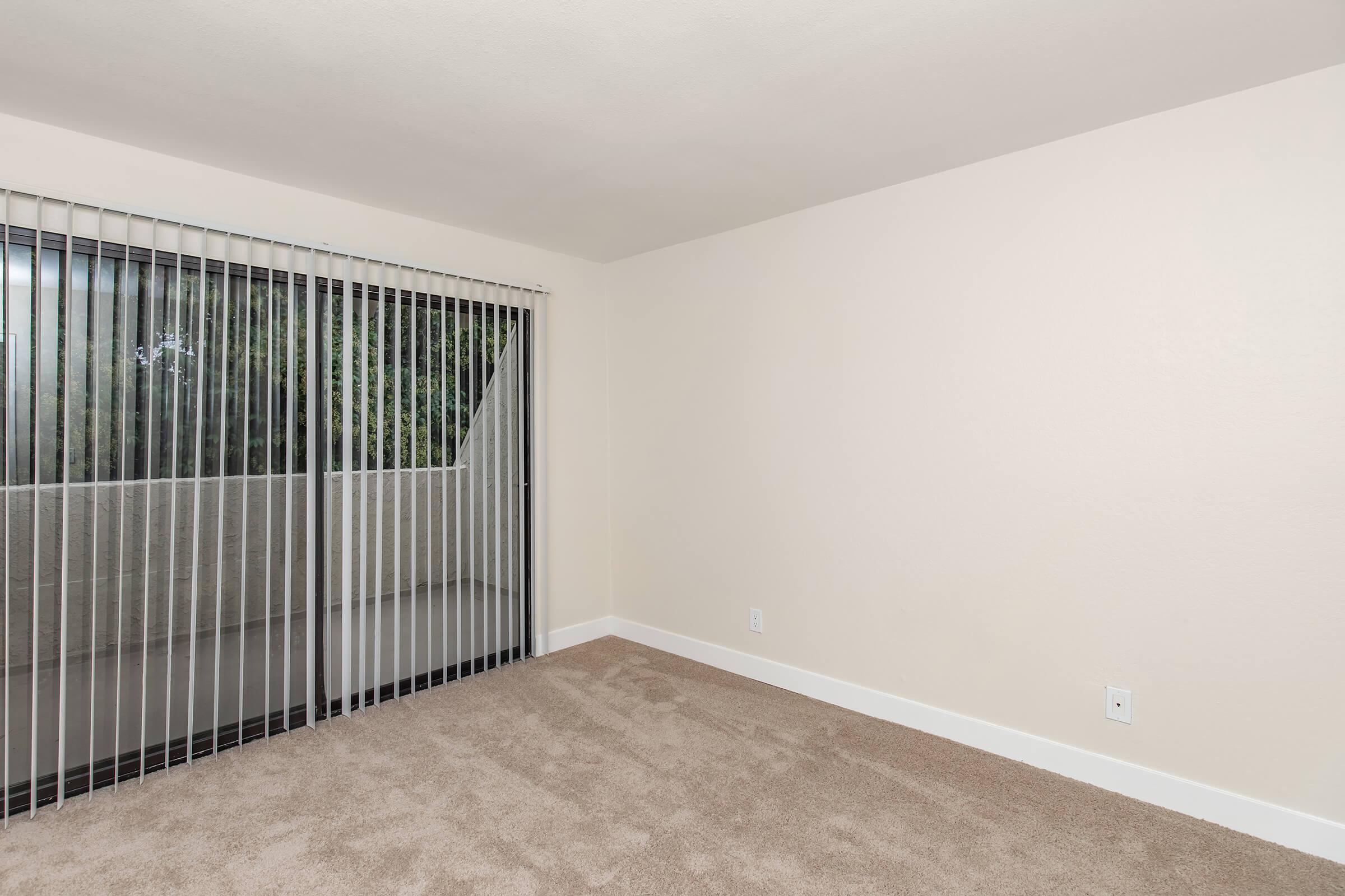 Empty room with beige walls and light brown carpet. There are vertical blinds covering a large sliding glass door that leads to a balcony. The room is well-lit and appears spacious, with no furniture or decorations.