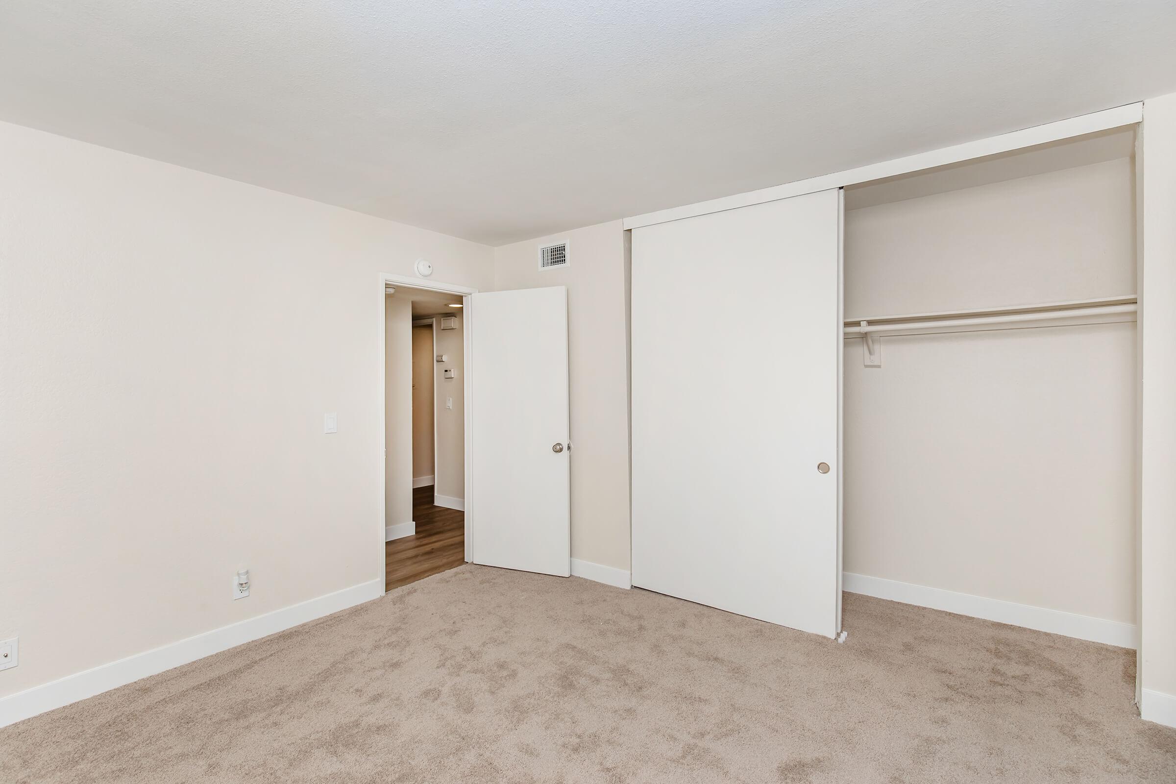 An empty room with beige walls and carpeting. A closet with sliding white doors is present on the right. An open doorway leads to another room in the background, suggesting a spacious layout. Natural light brightens the area, creating a clean and inviting atmosphere.