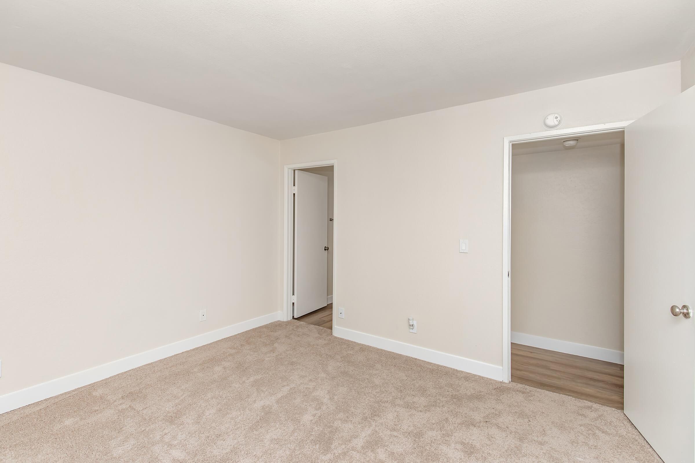 A spacious, empty room with light beige walls and soft, plush carpet. There are two doorways leading to adjacent areas, one partially open. Natural light brightens the room, creating a clean and inviting atmosphere. Perfect for a fresh decorating project or new living space.