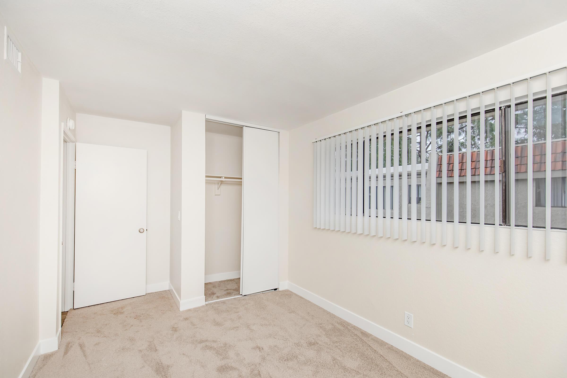 A clean, empty room featuring beige carpet, white walls, and vertical blinds on a window. There is a closet with sliding doors on one side and a simple white door leading to another space. Natural light enters through the window, creating a bright and airy atmosphere.