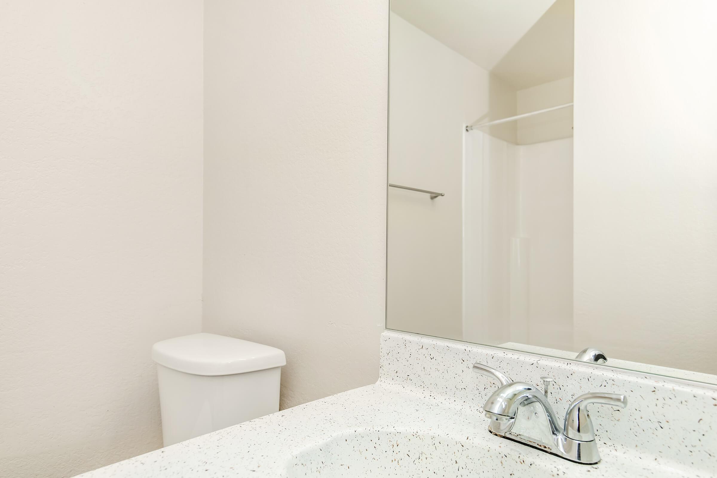 A clean bathroom interior featuring a white countertop with a chrome faucet, a large mirror reflecting the space, a white toilet, and a shower area with a glass door. The walls are painted in a light color, creating a bright and airy atmosphere.