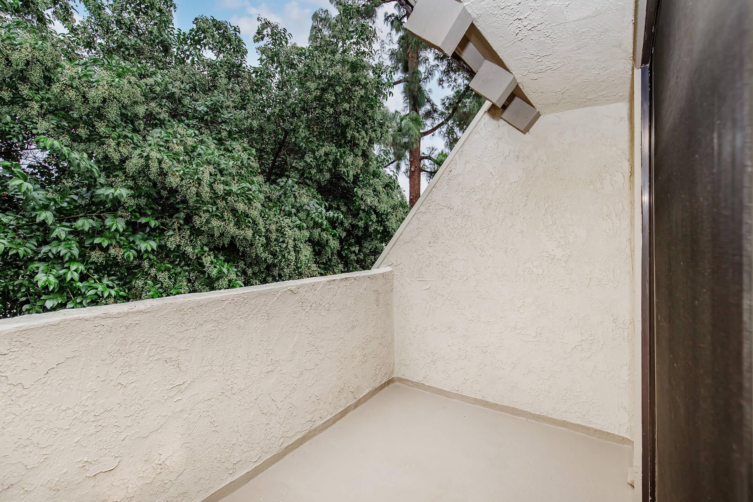 A spacious balcony with a textured white wall, overlooking a lush green landscape filled with trees. The area is well-lit, with an open sky visible in the background, providing a serene outdoor space.
