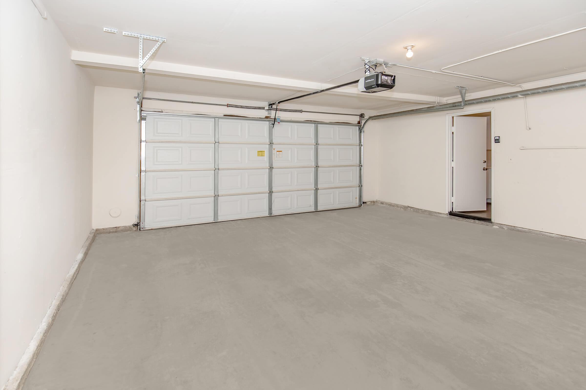 An empty garage with a smooth gray concrete floor, featuring a closed overhead door and a side door leading outside. The walls are painted white, and there is a ceiling light fixture illuminating the space. The garage appears clean and well-maintained, suitable for parking a vehicle or storage.