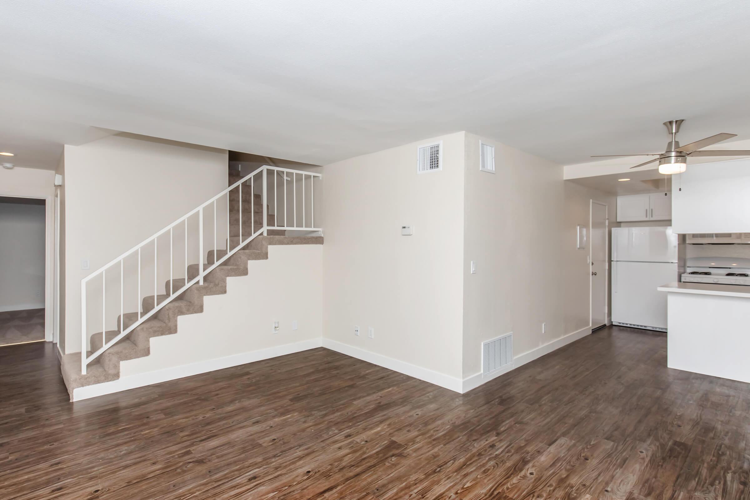 Interior view of a modern living space featuring a staircase leading to an upper level. The room has light-colored walls and dark wood flooring. A kitchen area is visible in the background, with a white refrigerator and ceiling fan enhancing the open layout.