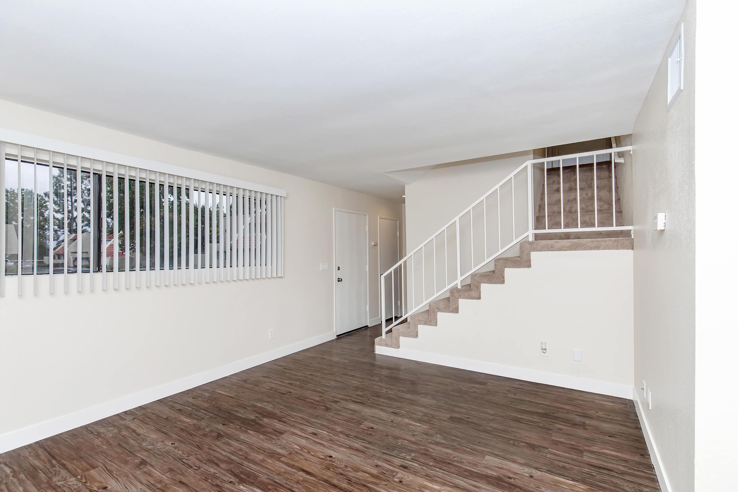 A spacious living area with light-colored walls and wooden flooring, featuring a large window with vertical blinds. A staircase with white railings leads to an upper level, while a door at the far end opens to the entrance. Natural light enhances the room’s bright and inviting atmosphere.