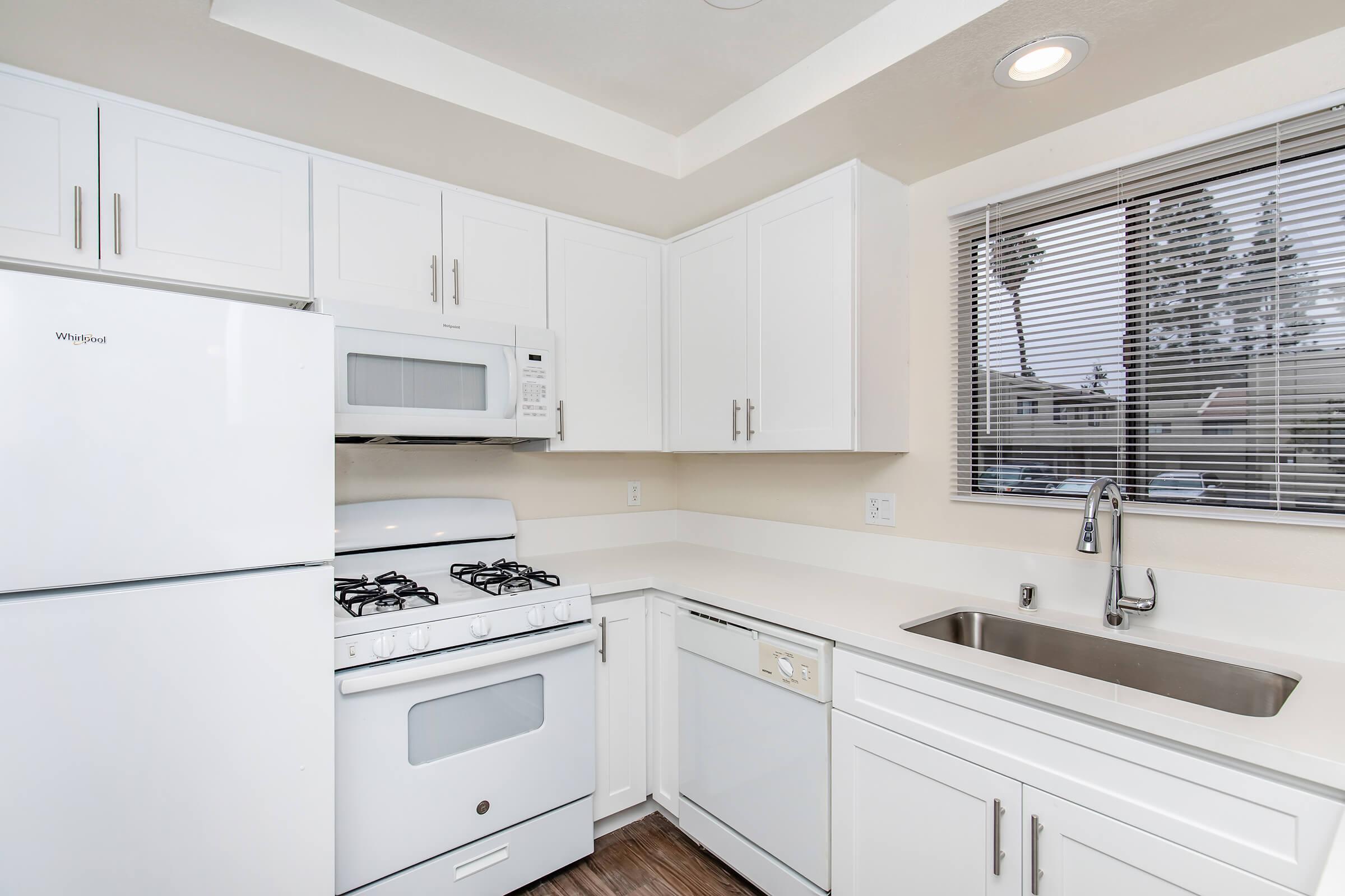 Modern kitchen featuring white cabinetry, a white refrigerator, a microwave above a gas stove, and a dishwasher. There's a sink under a window with blinds. The countertops are light-colored, and the flooring is hardwood. The overall design is bright and contemporary.