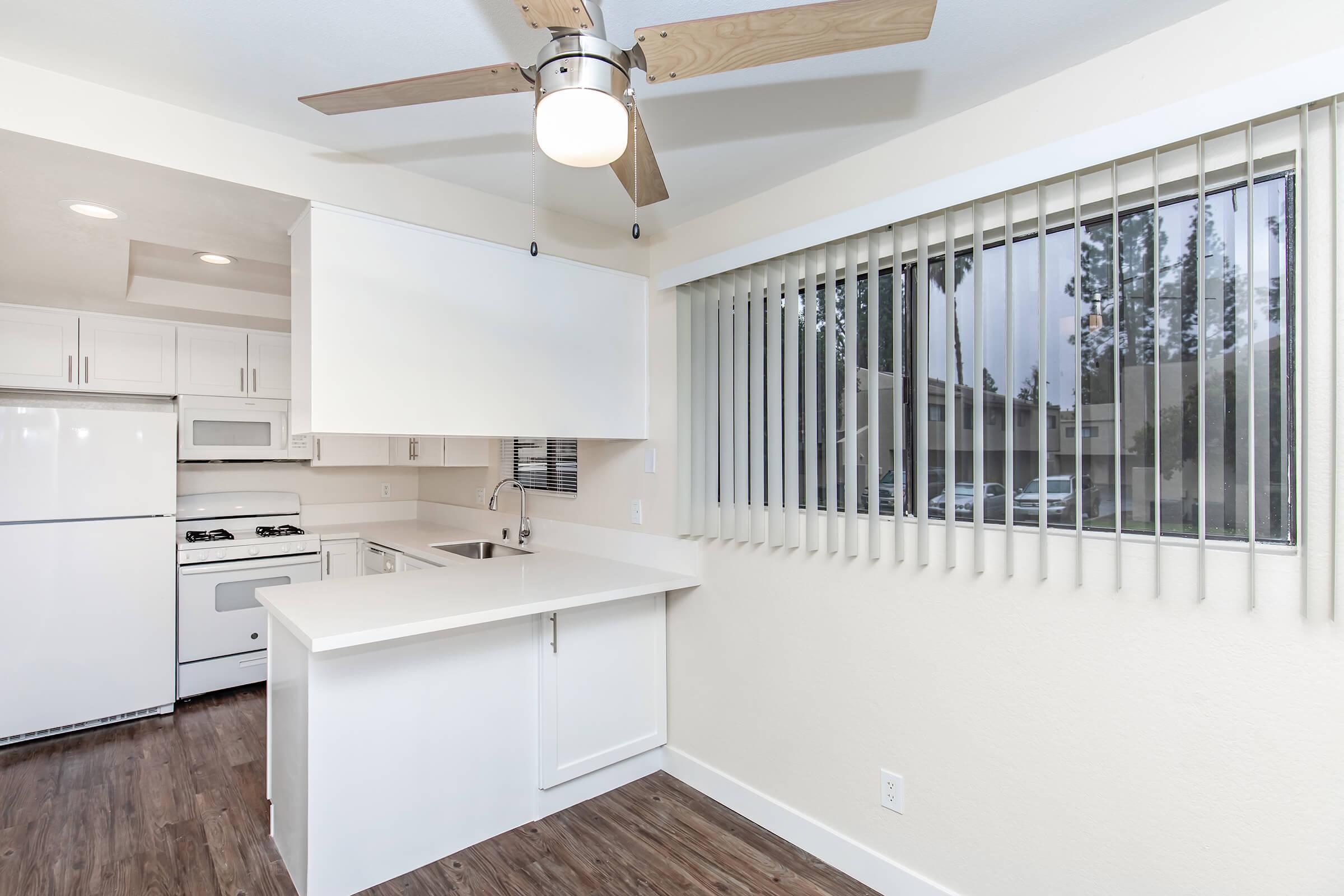 Bright kitchen with white cabinets and appliances, including a refrigerator and stove. Features a bar area with a countertop and a ceiling fan. Large window with blinds allows natural light, and a view of an exterior area. Modern design with wooden flooring.