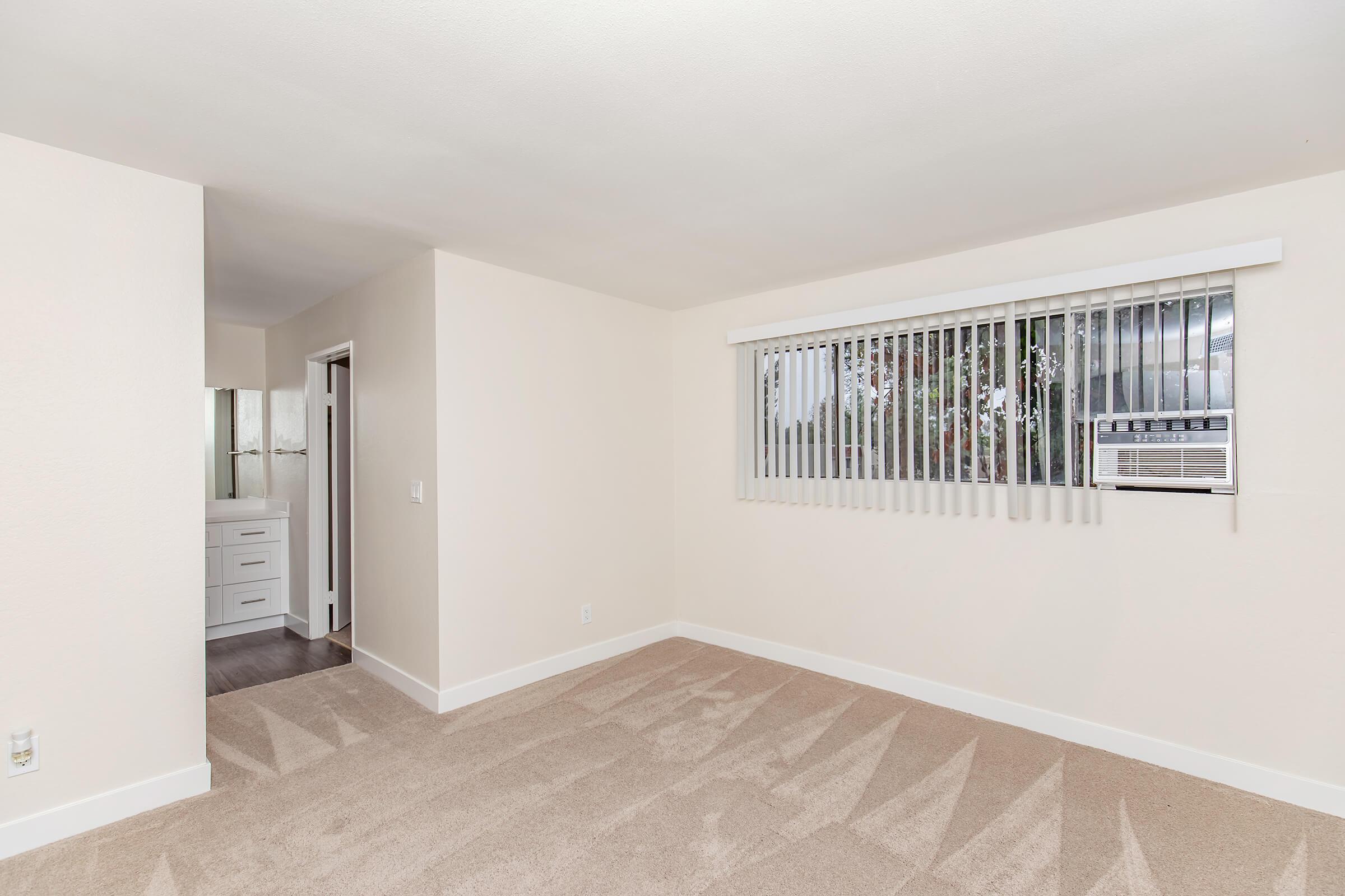 A bright and spacious room with beige walls and carpet. A window with vertical blinds lets in natural light; an air conditioning unit is visible above the window. In the background, a doorway leads to a bathroom area with white cabinets. The overall atmosphere is clean and inviting.