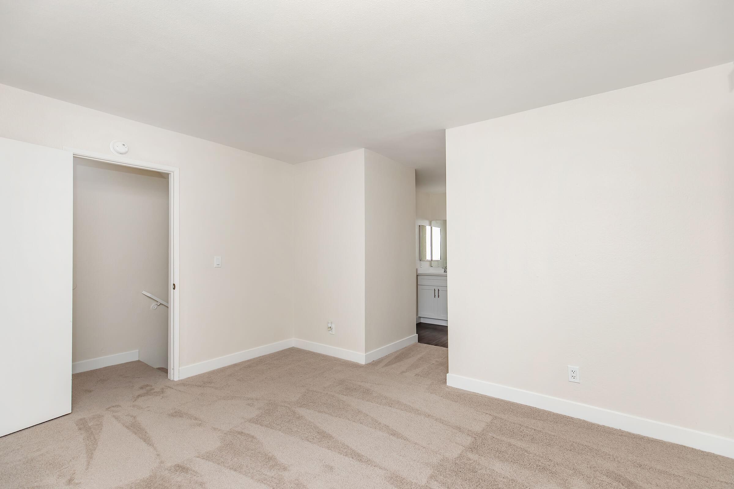 Empty room with light-colored walls and beige carpet. A door on the left leads to another space, while the room opens towards a small hallway. In the background, a bathroom area is visible with light fixtures reflecting in a mirror. The overall atmosphere is bright and spacious.
