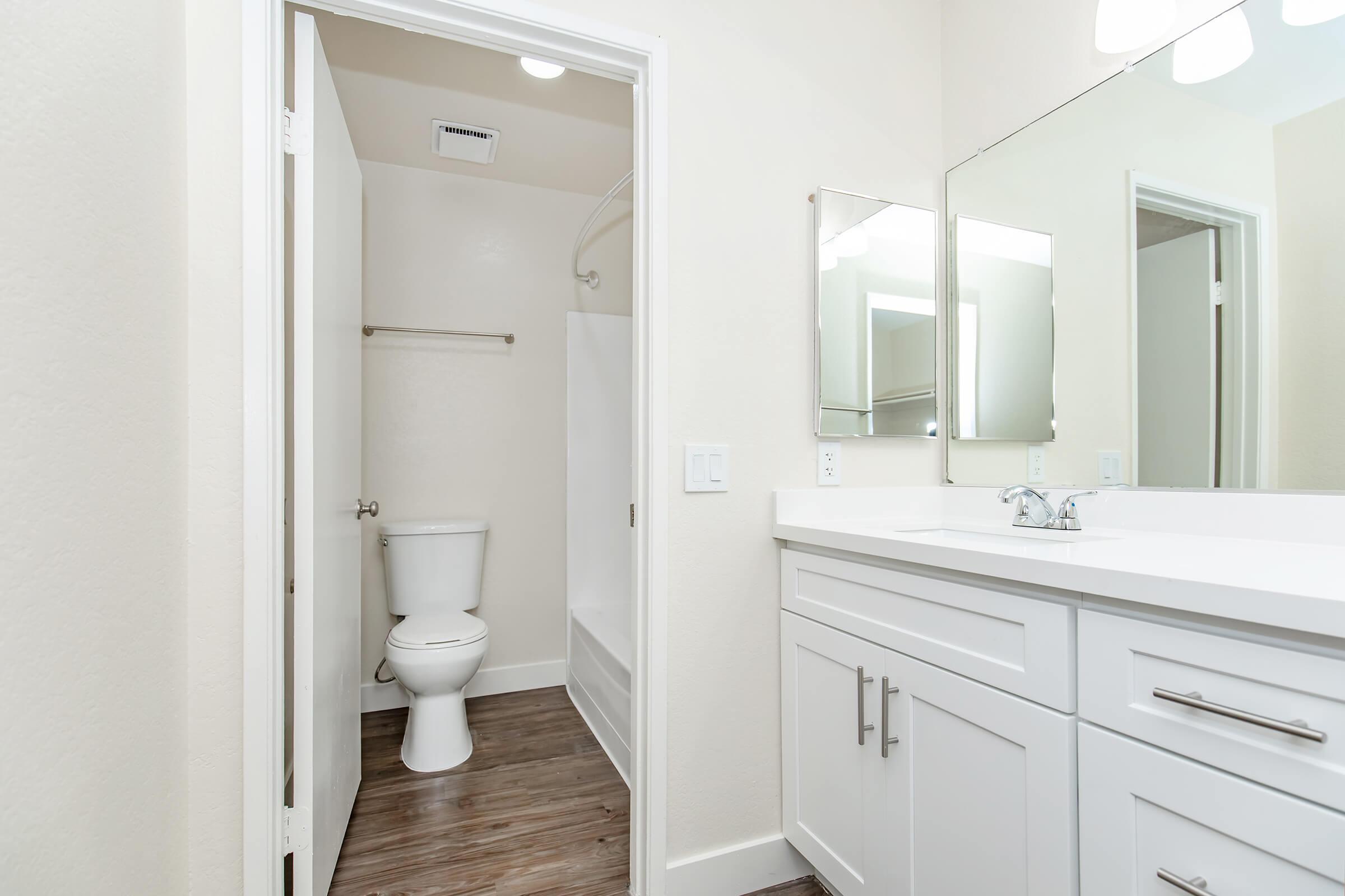 A modern bathroom featuring a white vanity with a sink, a large mirror above, a toilet in a separate area, and a shower with a clear glass door. The walls are painted light beige, and the flooring is a dark wood laminate. Bright overhead lighting enhances the clean, spacious look.