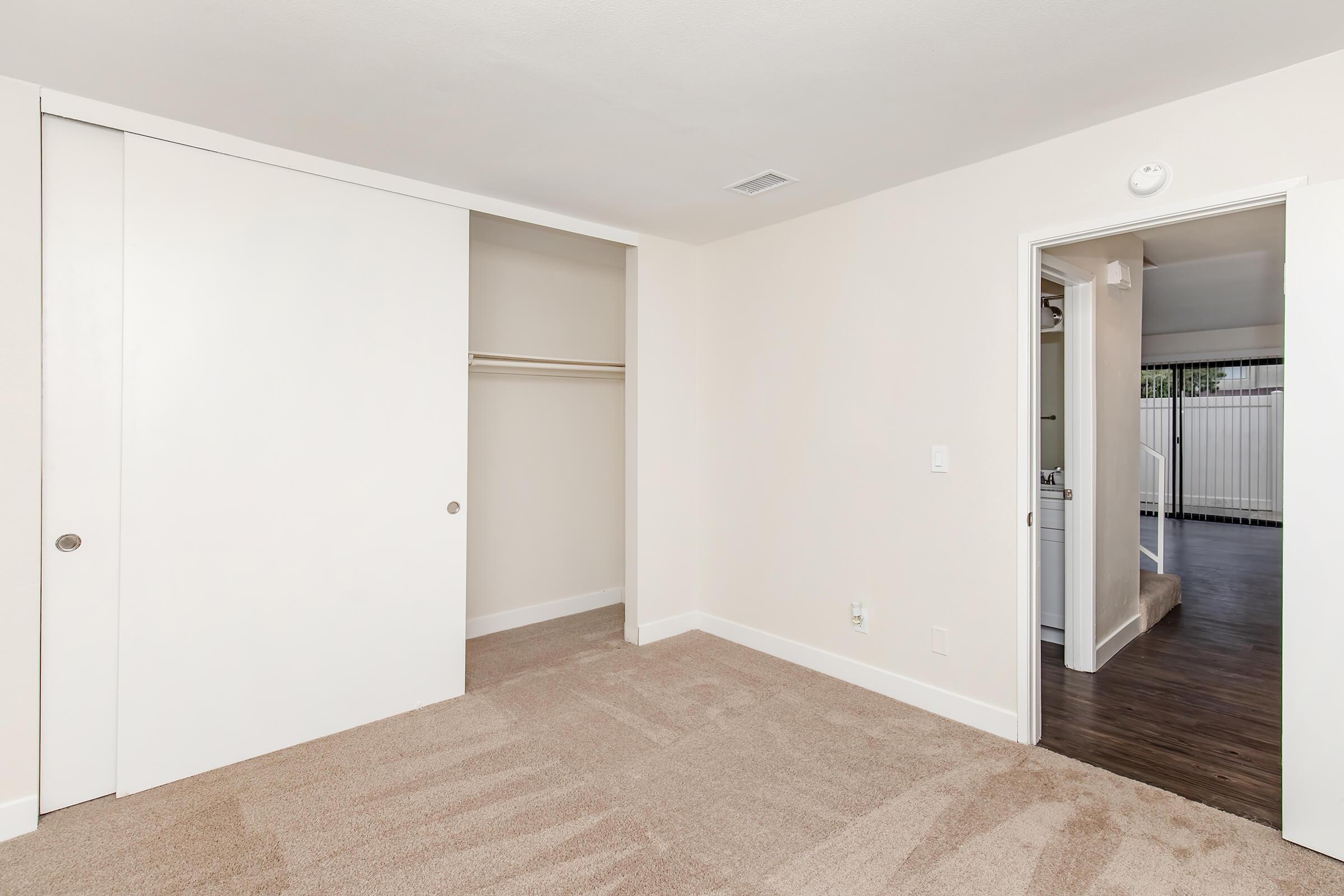 Interior view of a room featuring beige walls and carpet. A closet with sliding white doors is visible on the left, while a doorway leads to another area on the right. The room is well-lit, showcasing a clean and minimalist design.