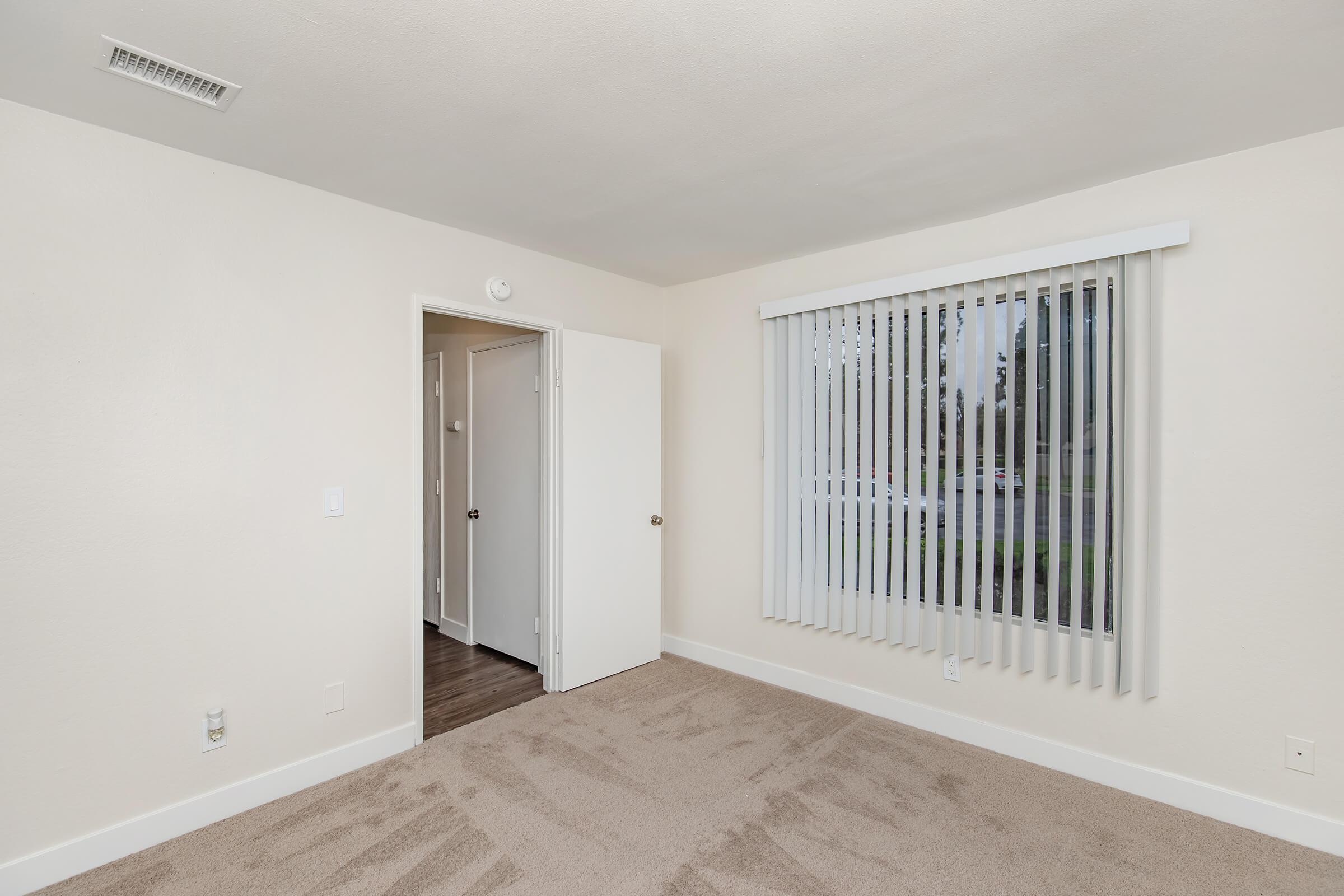 A vacant bedroom featuring beige carpet, light-colored walls, and a large window with vertical blinds. An open door leads to another room, and there's a ceiling vent. The space is well-lit and minimalistic, creating a neutral and inviting atmosphere.