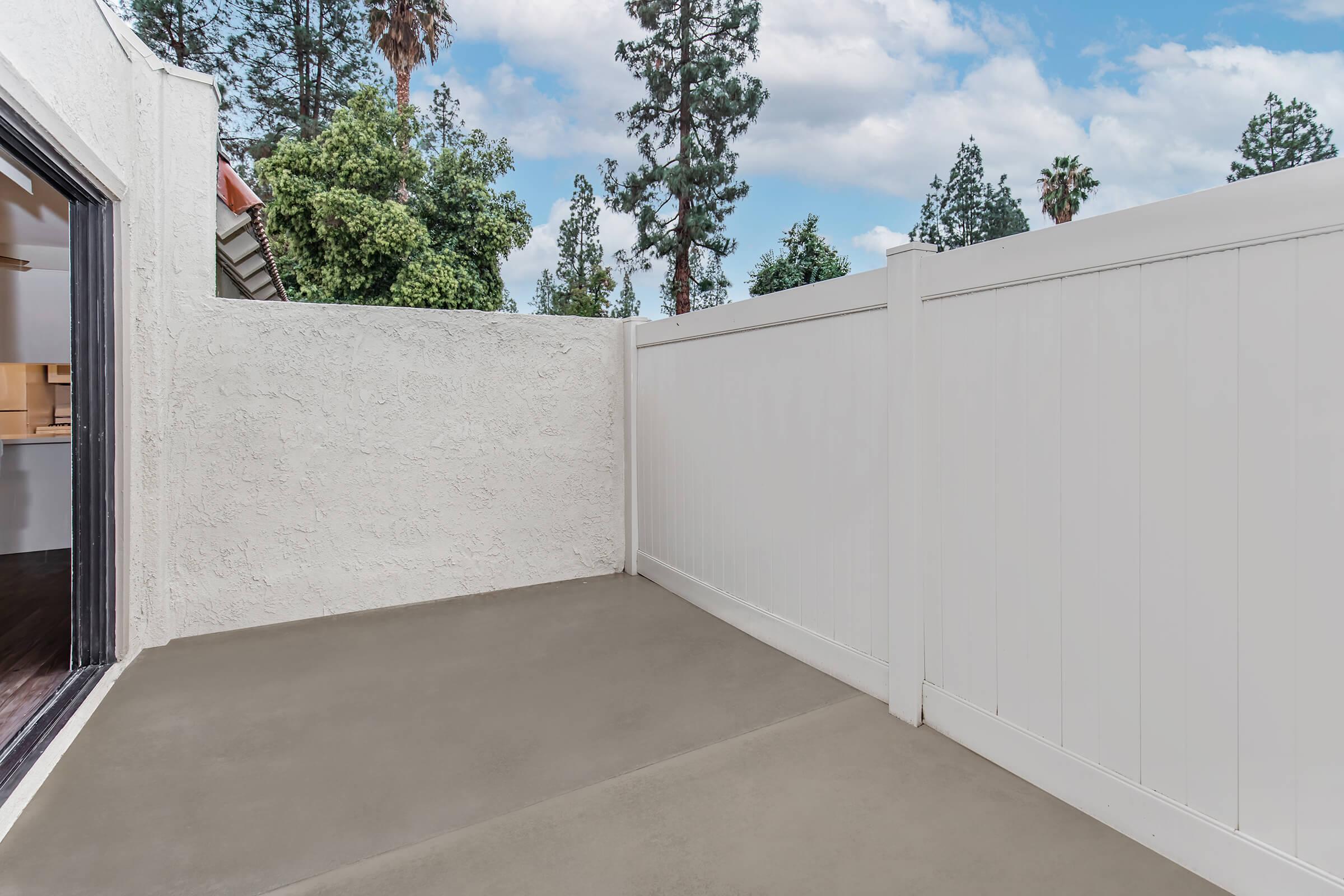 A small outdoor patio area with a concrete floor, enclosed by a white fence. Visible greenery and tall trees in the background, along with a partially open doorway leading inside. The sky is partly cloudy.