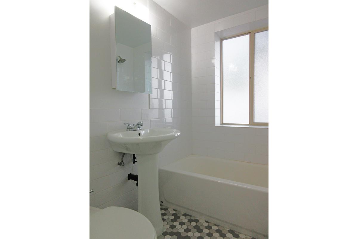 A modern bathroom featuring a white tiled wall, a bathtub, a pedestal sink with a mirror above, and a window allowing natural light. The flooring is made of hexagonal patterned tiles, creating a clean and minimalist aesthetic.