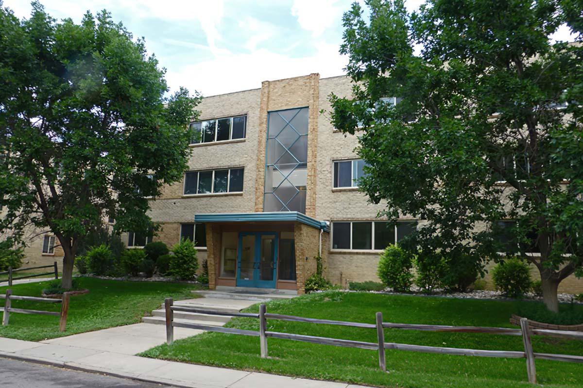 A three-story brick apartment building with a central entrance featuring a blue awning. Grassy areas and small trees surround the building, along with a wooden fence in the foreground. The sky is partly cloudy, creating a bright atmosphere.