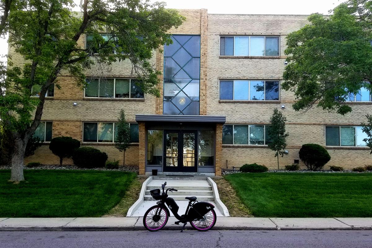 A modern brick apartment building with large windows and greenery in front. A bicycle is parked on the sidewalk in the foreground, with neatly trimmed bushes and trees lining the entrance path. The scene is well-lit, suggesting a pleasant day.