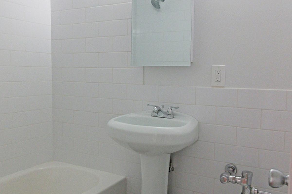 A clean, minimalistic bathroom featuring a white tiled wall, a bathtub, a pedestal sink with modern faucets, and a wall-mounted mirror. The space is well-lit and maintained, emphasizing simplicity and functionality in its design.