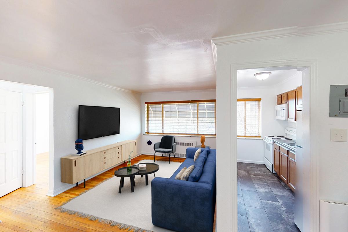 A bright and modern living room featuring a blue couch, a flat-screen TV mounted on the wall, a light wood TV stand, and a small black chair. Natural light streams through windows with wooden blinds, and the room connects to a kitchen area with white cabinets and appliances, all set on a dark tile floor.
