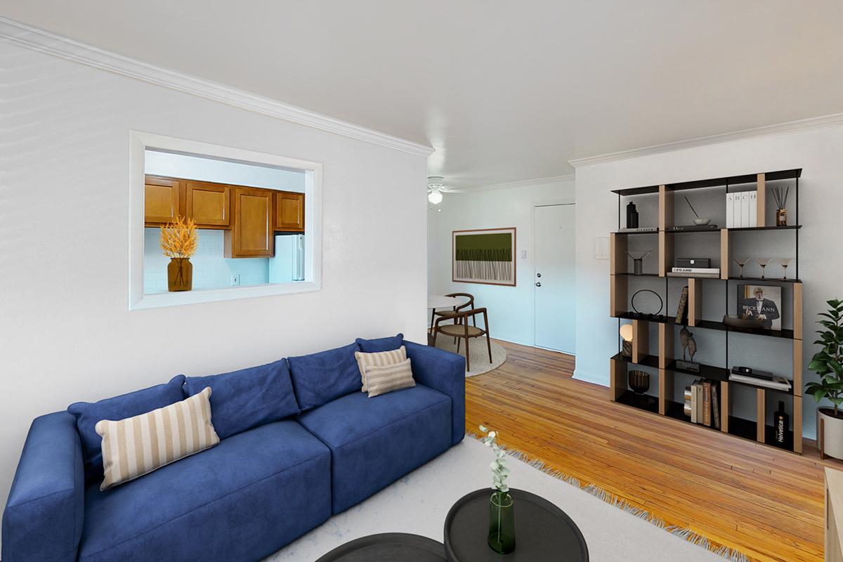 A modern living room featuring a blue sofa with striped cushions, a black coffee table, and a stylish bookshelf filled with decor. Light wood floors and white walls create a bright atmosphere. An open wall connects to a kitchen area with wooden cabinets. A decorative plant adds a touch of greenery.