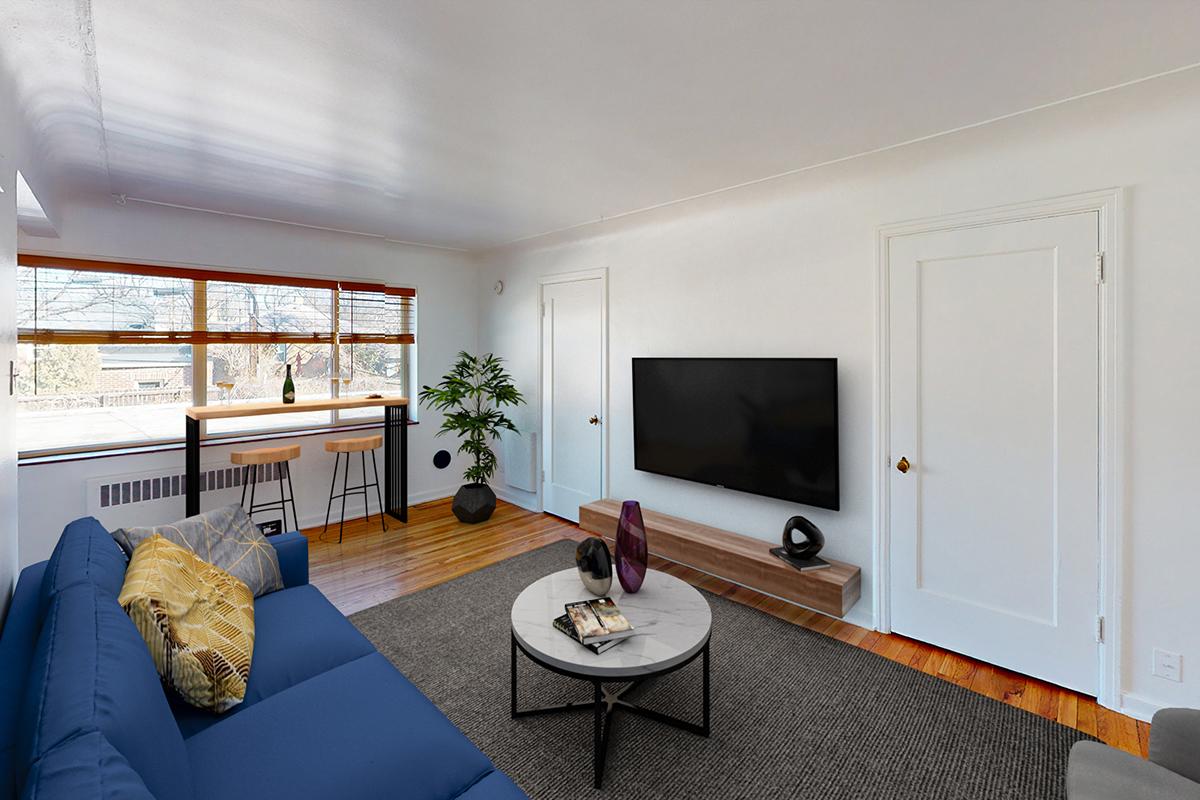 A cozy living room featuring a blue sofa with decorative pillows, a round coffee table on a rug, and a sleek TV mounted on the wall. A small dining area with a barstool setup is visible, alongside a green indoor plant and large windows allowing natural light to fill the space.