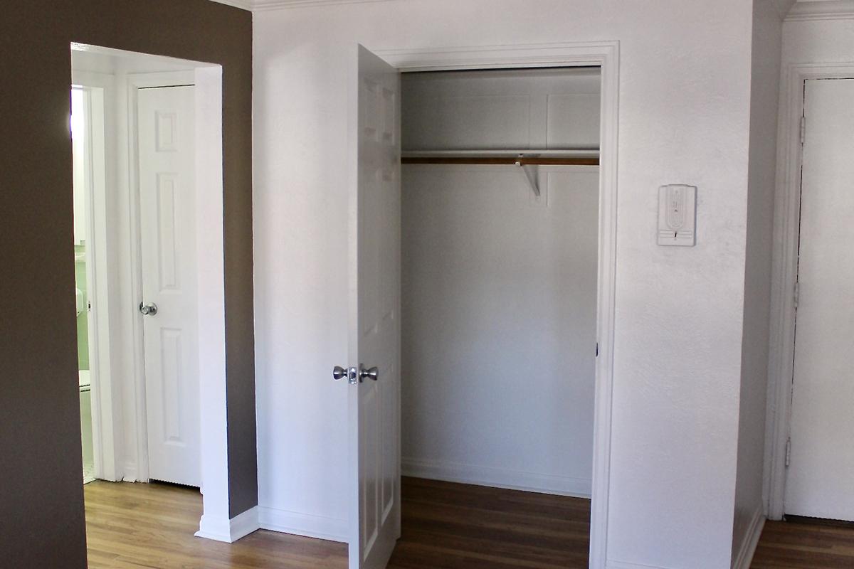 A well-lit, empty room featuring two doorways leading to other spaces. One doorway opens to a closet with a wooden rod for hanging clothes. The walls are painted in neutral colors, and the flooring is wooden. The room has a clean and spacious appearance, suitable for various uses.