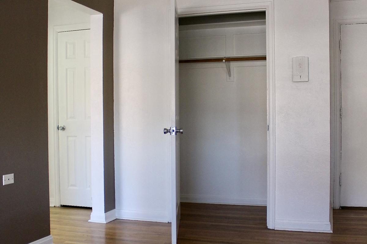 A minimalistic interior of a room featuring open doors leading to a closet and another room. The walls are painted in two colors: one side is brown and the other white. The wooden floor is visible, and the closet is empty with a single bar for hanging clothes.