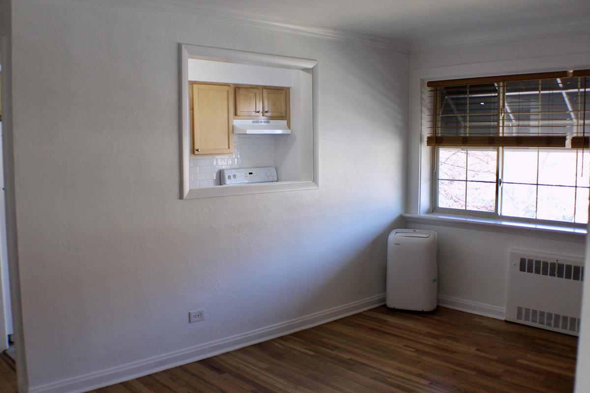 A bright, empty room with wooden flooring and plain white walls. There is a window with wooden blinds letting in light, and an air conditioning unit on the wall. A small open space leads to a kitchen with wooden cabinets behind a white frame.