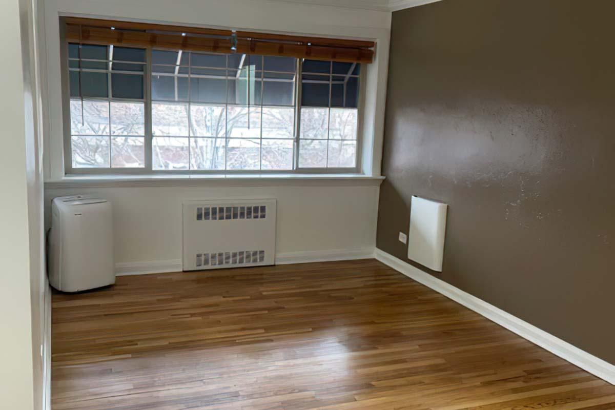 A small, empty room featuring hardwood flooring, a large window with blinds, and a beige wall. An air conditioning unit is positioned in one corner, and a radiator is visible on the wall beneath the window. The overall atmosphere is bright and minimalistic.