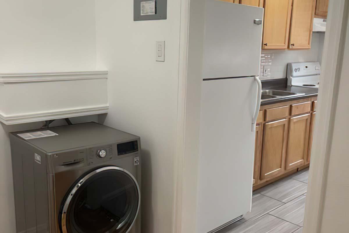 A small laundry area featuring a modern washing machine next to a white refrigerator. The space includes wooden cabinets and a kitchen counter with a sink, creating a tidy and functional home environment. The flooring is tiled, enhancing the overall appearance.