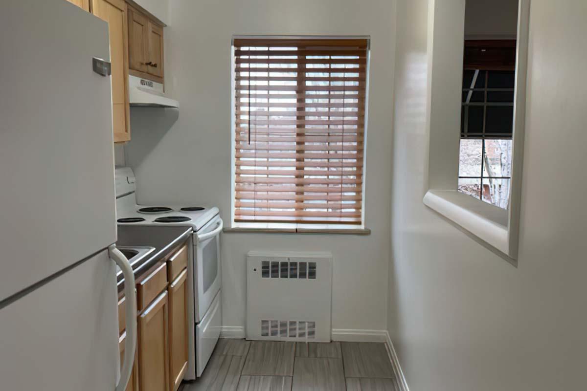 A small kitchen featuring white appliances, including a refrigerator and stove, with light wood cabinets. There's a window with wooden blinds, allowing natural light, and tile flooring. An extra heater unit is visible on the wall below the window. The overall space is clean and well-lit.