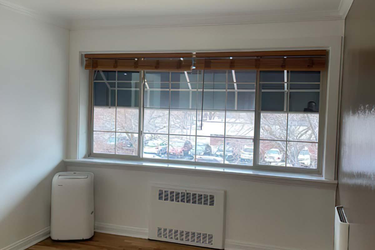 A bright room with a window featuring bamboo blinds. Through the window, a view of a parking lot with several cars is visible. The room has light-colored walls and wooden flooring, with an air conditioning unit beneath the window. Sunlight filters in, creating a warm atmosphere.