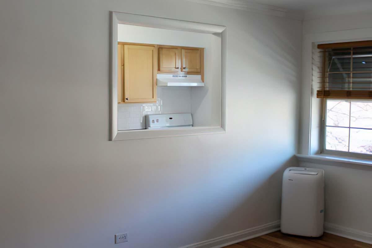 A bright, empty room featuring a large, framed opening that connects to a kitchen area with wooden cabinets and a white backsplash. Natural light filters in through a window with blinds, and a white air conditioning unit is positioned in the corner, enhancing the airy feel of the space.