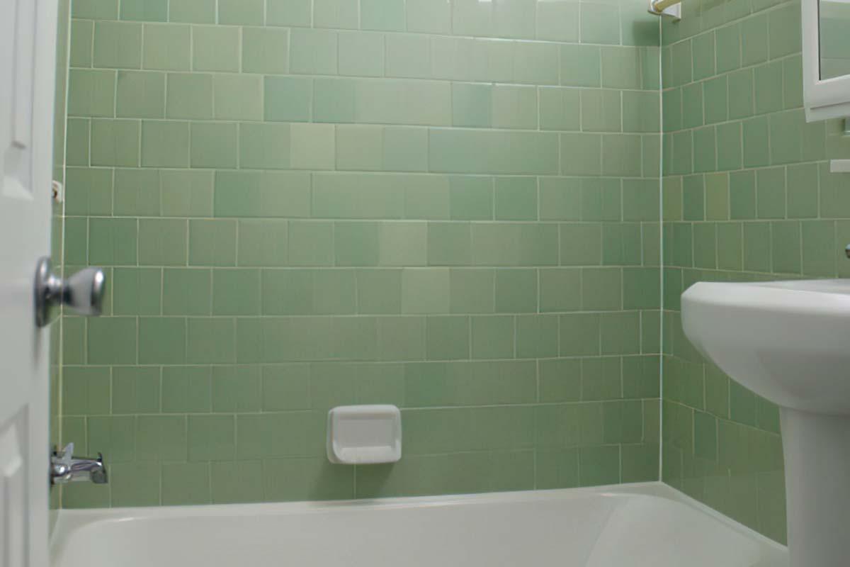 A bathroom with green tiled walls, featuring a white bathtub and a toilet. A mirror is mounted on the wall above the sink, and there is a small shelf. The overall atmosphere is clean and minimalist, with a focus on the green tile design.