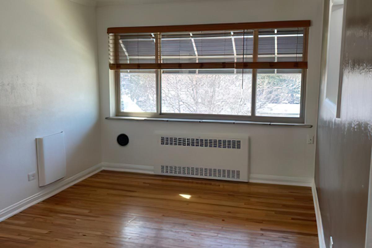 A bright, empty room with hardwood flooring and a large window adorned with wooden blinds. The window provides natural light, and there is a heater beneath it. A small circular vent is visible on the wall. The overall space has a clean, minimalist appearance.