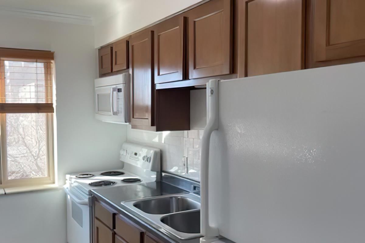 A modern kitchen featuring wooden cabinetry, a white refrigerator, a stove and oven, a microwave mounted above the range, and a double sink. Natural light streams in through a window with blinds. The countertops are clean and the space is well-organized.