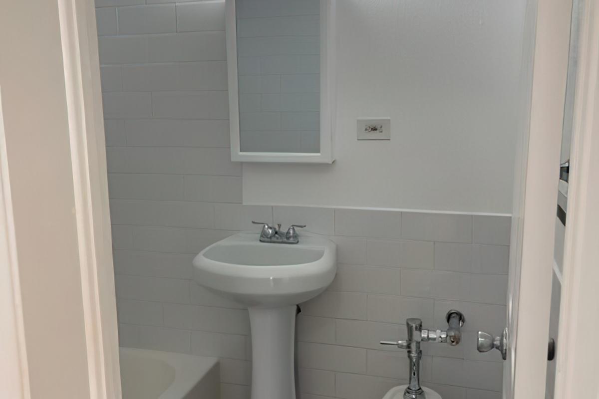 A clean, modern bathroom featuring a white pedestal sink, a wall-mounted mirror, and white tiled walls. In the background, there's a bathtub and a toilet. The overall aesthetic is minimalistic and bright, with natural light illuminating the space.