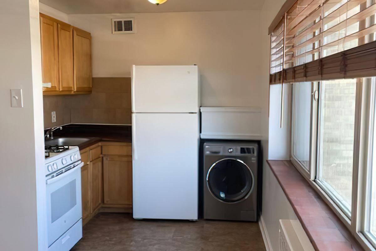 A small kitchen featuring wooden cabinets, a white refrigerator, a washing machine, and a stove. Natural light comes through a window with blinds, illuminating the room. The floor is tiled, and there is a countertop next to the appliances.