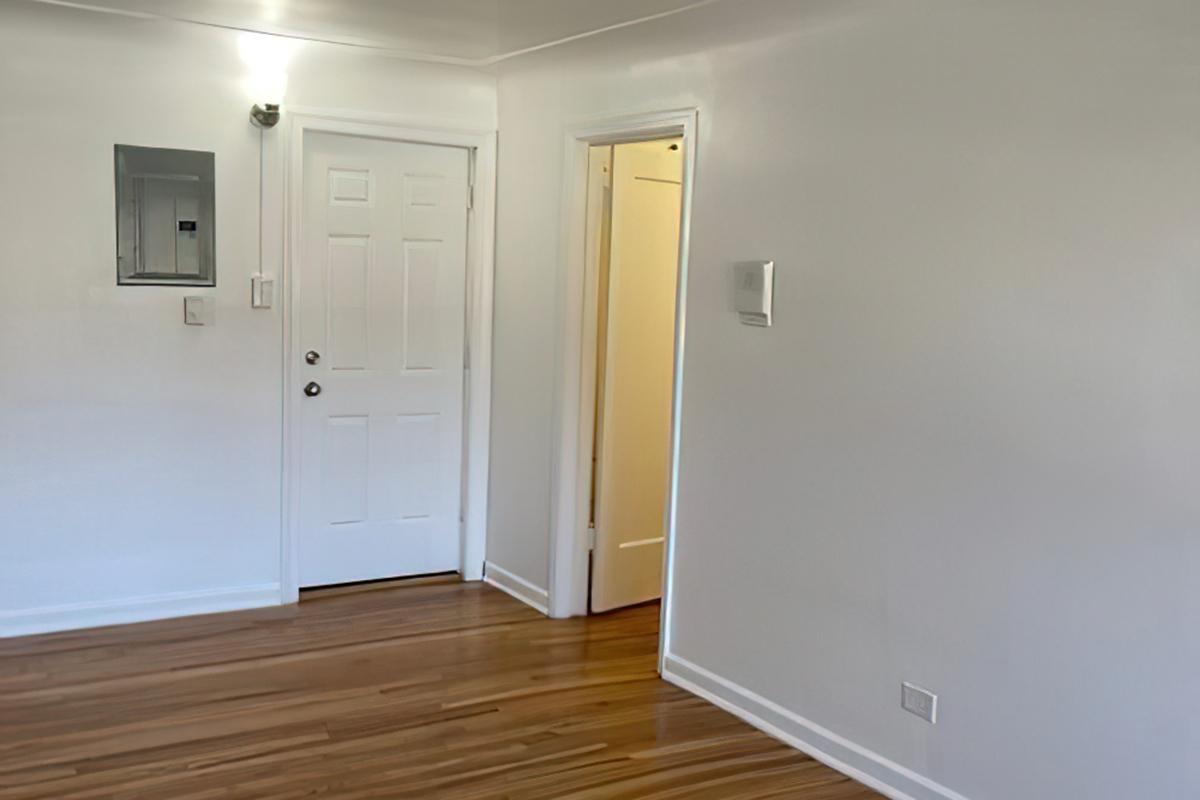 A brightly lit room featuring light-colored walls and polished wood flooring. There is a white door on the left leading outside, a closed door on the right, and a small electrical panel on the wall. The overall appearance is clean and uncluttered, suggesting a modern living space.