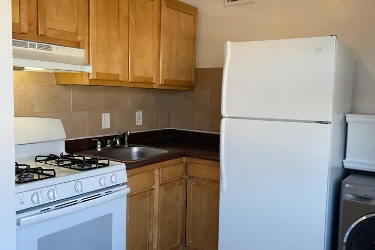 A small kitchen featuring wooden cabinets, a white stove with a gas cooktop, a white refrigerator, a sink with a faucet, and a beige tiled backsplash. A washing machine is visible in the corner. The space is bright and functional, designed for everyday cooking and laundry needs.