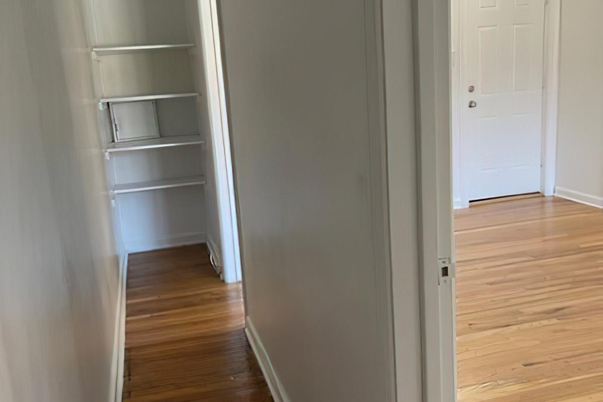 A view of a light-colored hallway featuring smooth, painted walls and hardwood flooring. On the left, there is a shelf unit built into the wall. At the end of the hallway, a door opens to another room, which is brightly lit and has a white door visible.