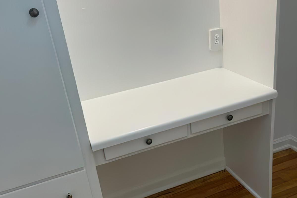 A clean white desk with two drawers, positioned against a wall. The surface is smooth and minimalist, with an electrical outlet visible on the wall beside it. The surrounding area is well-lit, highlighting the simplicity of the design. The floor features wooden panels.