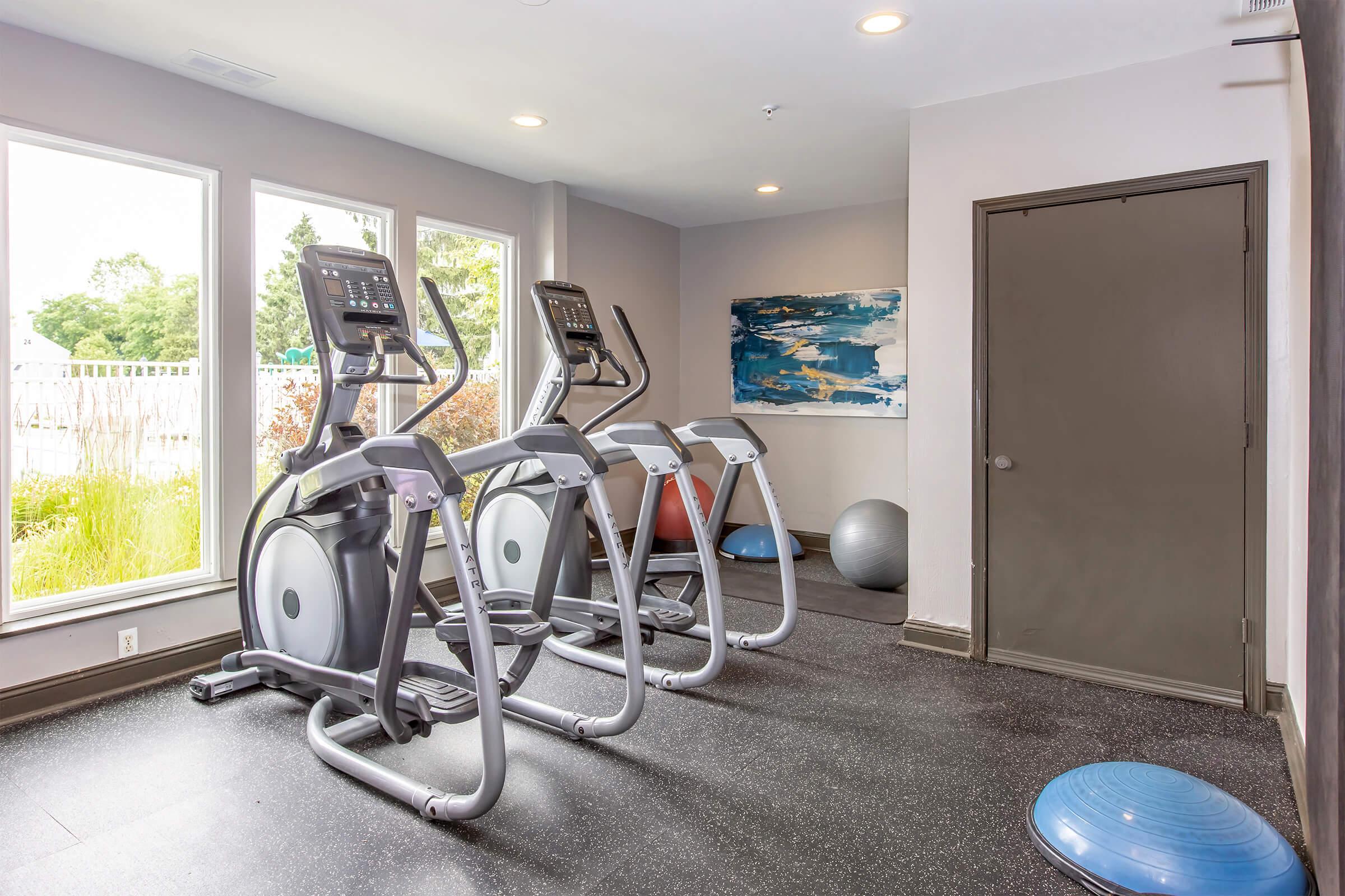 A modern fitness room featuring three elliptical machines facing large windows with natural light. The space includes a blue exercise ball and a balance disc on the floor, with minimalist decor and light-colored walls.
