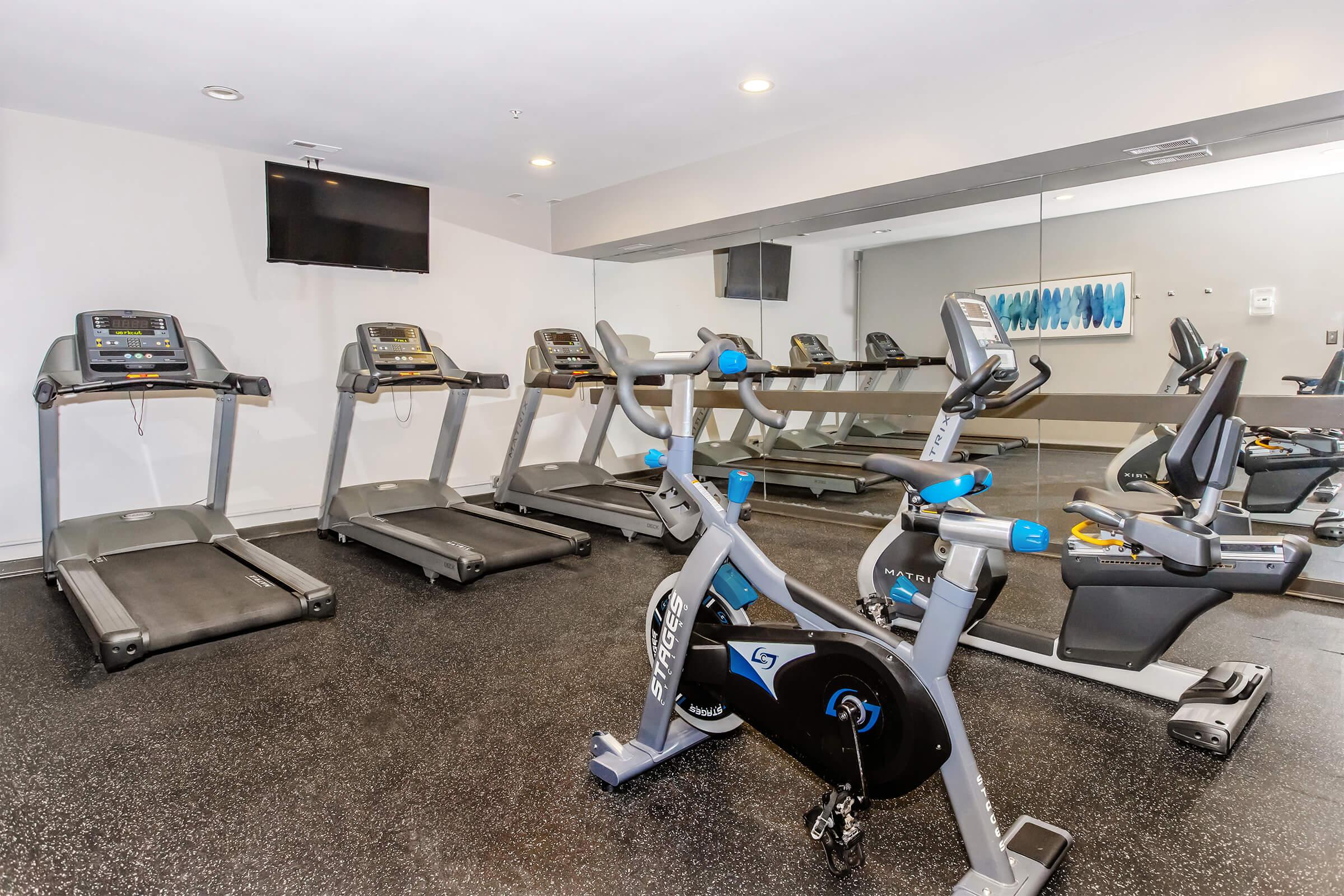A modern gym interior featuring four treadmills and a stationary bike, all equipped with digital displays. The floor is dark and textured, while a large mirror reflects the equipment. A flat-screen TV is mounted on the wall, and a piece of abstract art hangs nearby, adding a touch of color to the space.