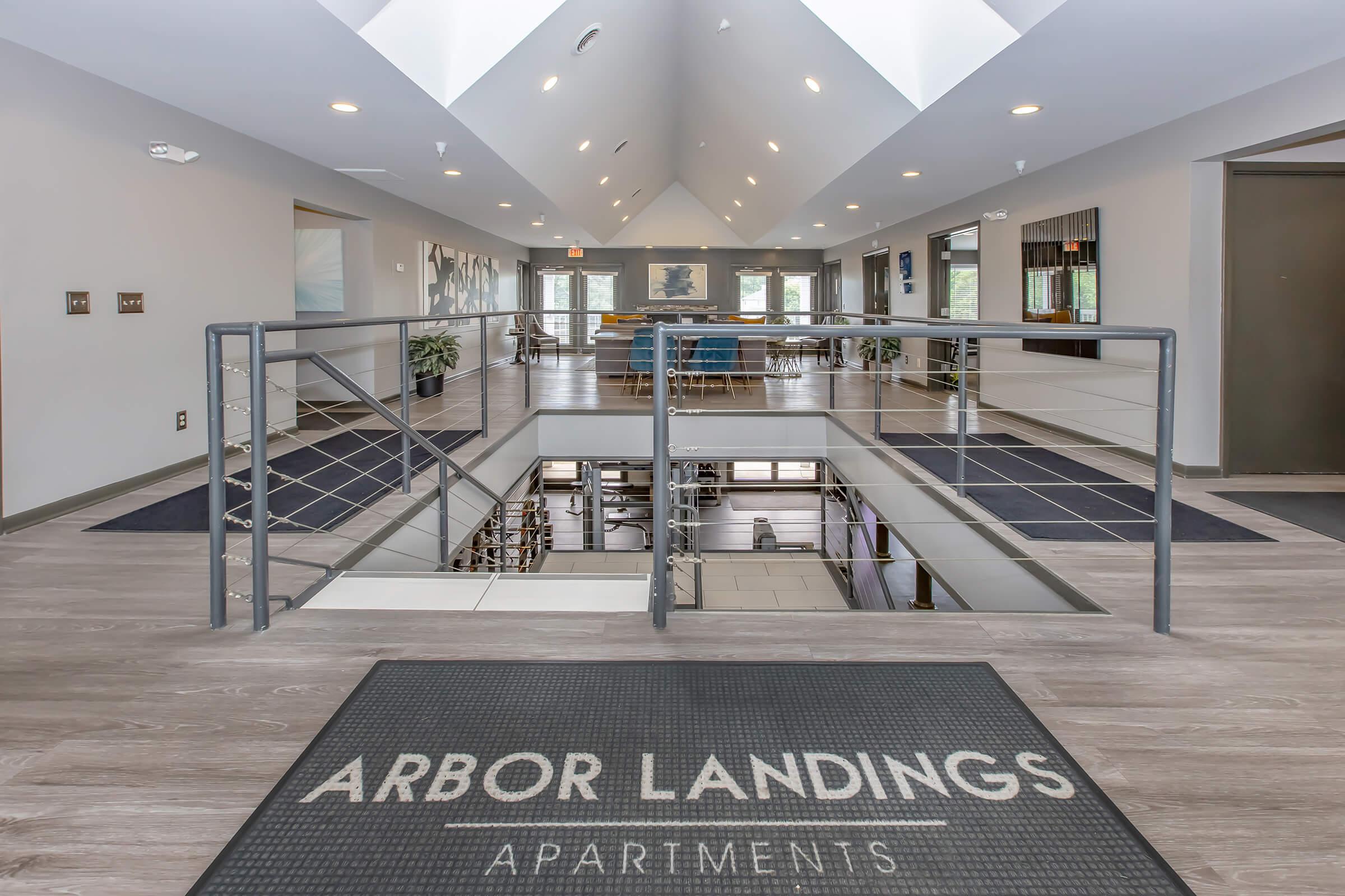 A modern apartment building lobby featuring a spacious, open layout with natural lighting. The entrance includes a decorative mat with "ARBOR LANDINGS APARTMENTS" printed on it. Stylish seating areas and artwork are visible in the background, enhancing the contemporary aesthetic.