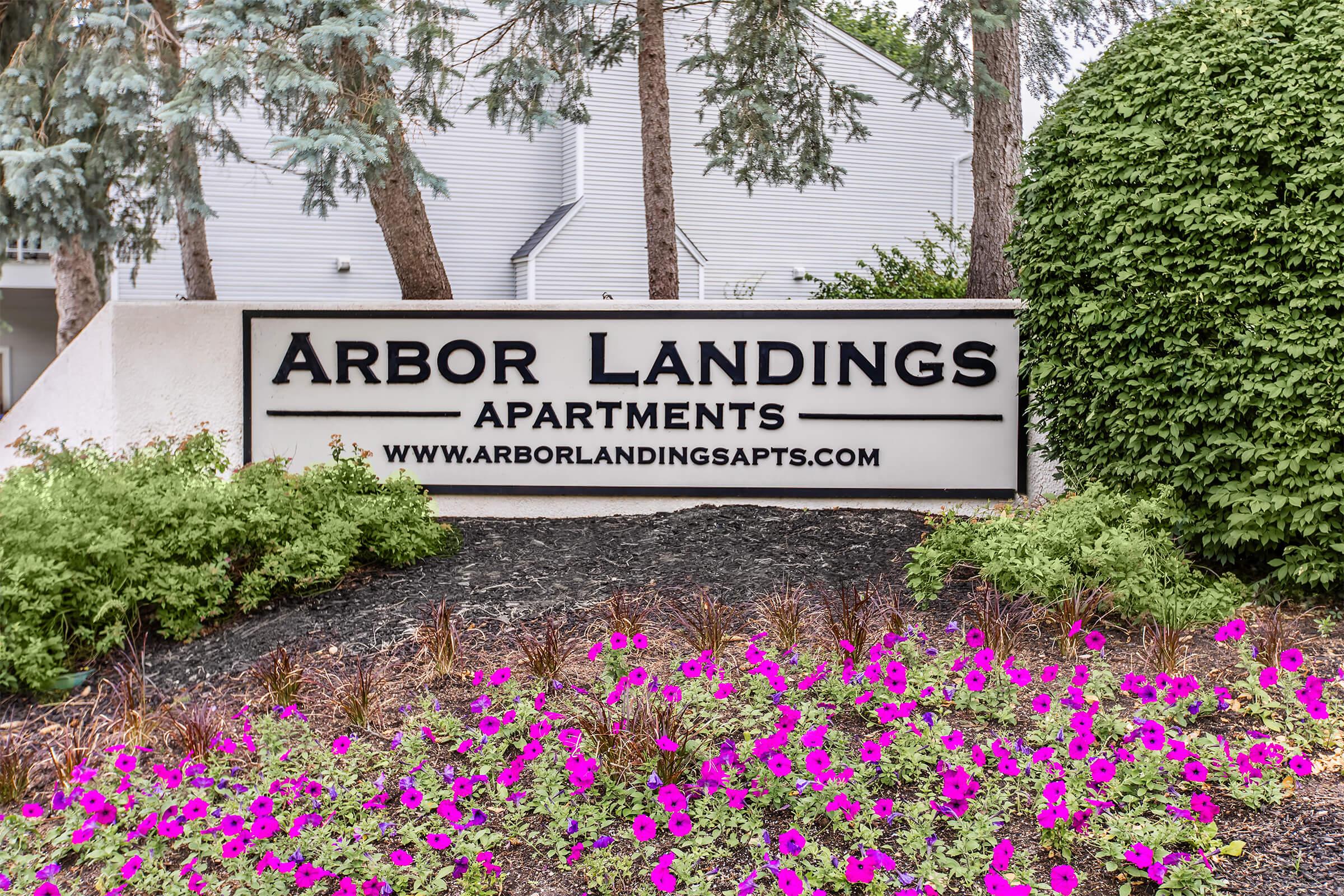 Sign for Arbor Landings Apartments, featuring the name prominently displayed. Surrounding the sign are well-maintained flower beds with vibrant pink and purple flowers, and greenery that adds to the inviting atmosphere. The background includes trees and a residential building.