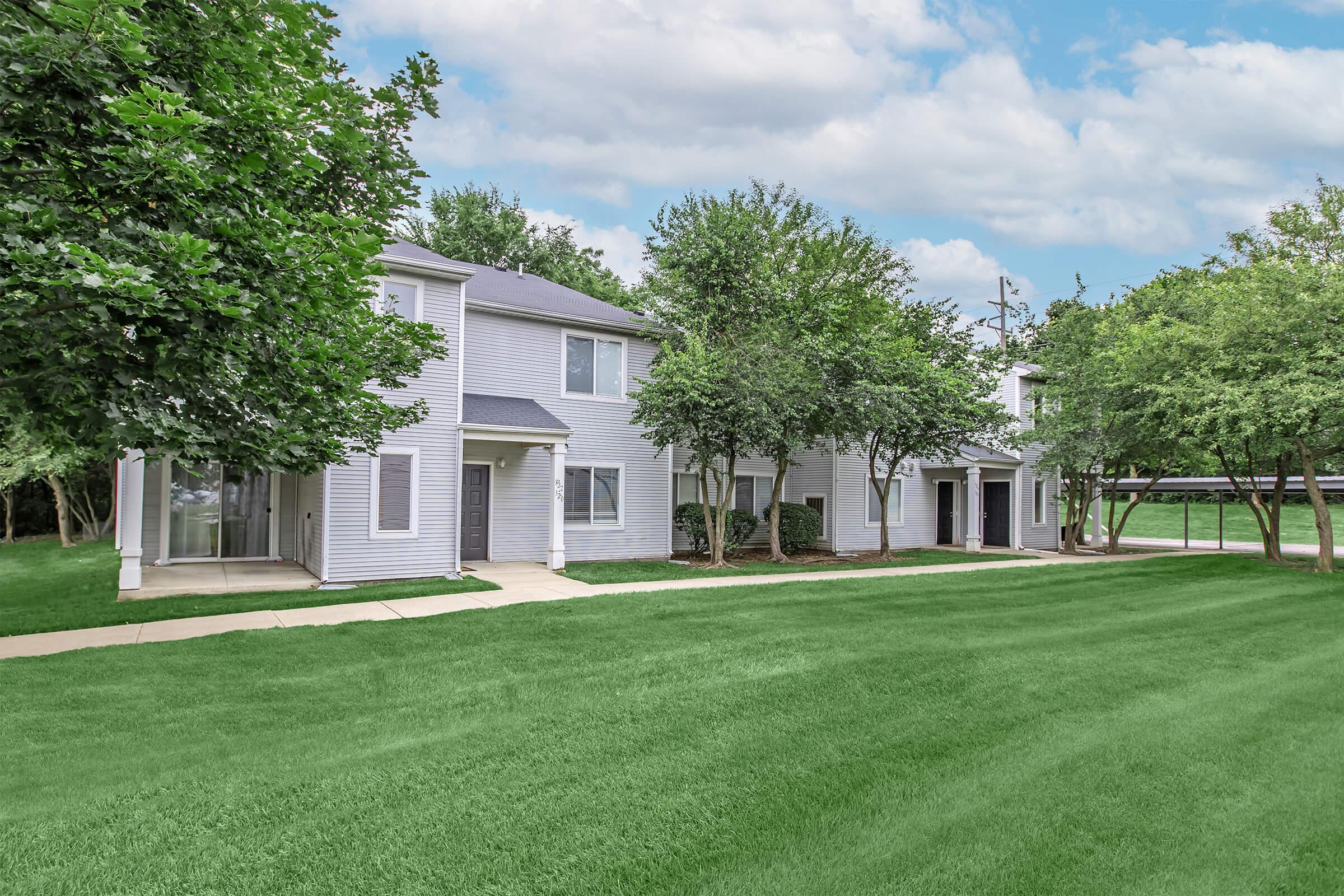 A landscaped exterior view of a two-story residential building surrounded by green grass and trees. The building features a light-colored facade, with several windows and a pathway leading to the entrance. The sky is partly cloudy, creating a bright atmosphere.