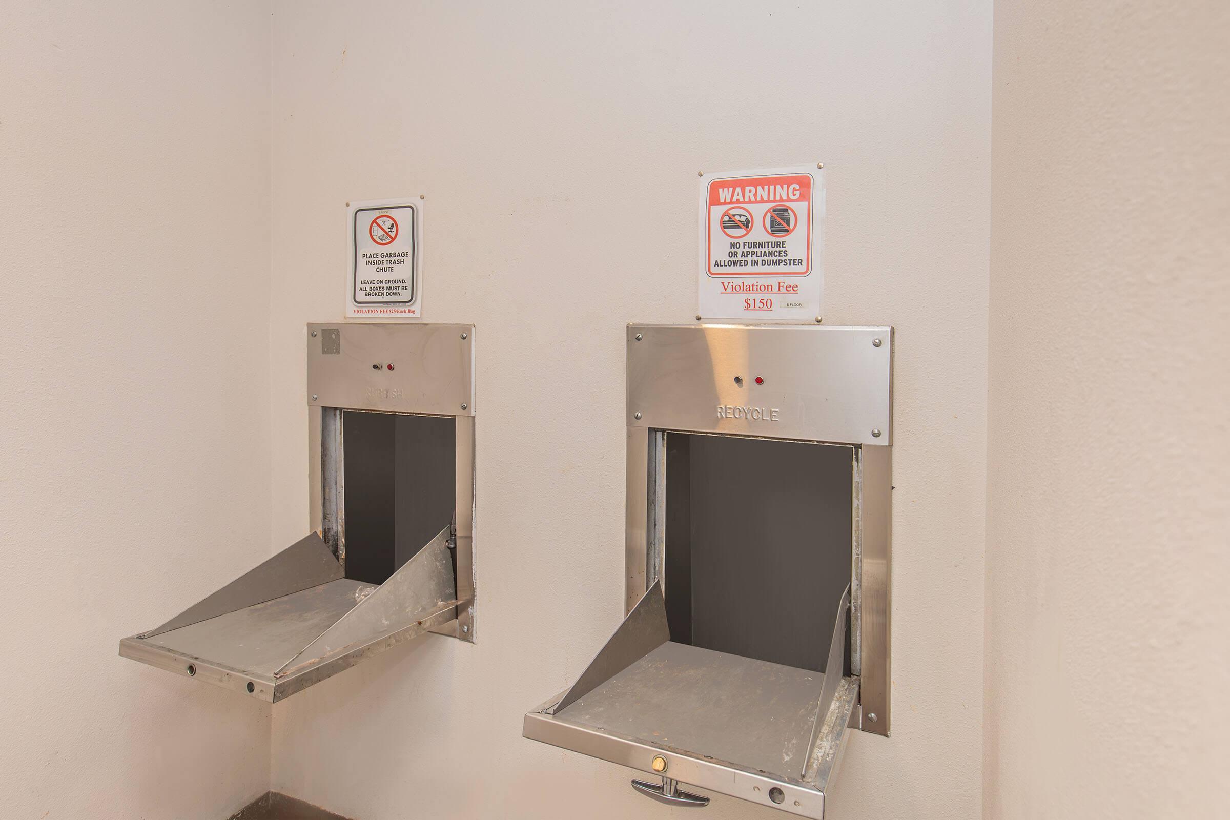 Two metal mail slots mounted on a wall, one above the other. Each slot has a small sign above it with warnings and text indicating a violation fee of $129. The wall is plain and light-colored, and the slots appear to be well-maintained.