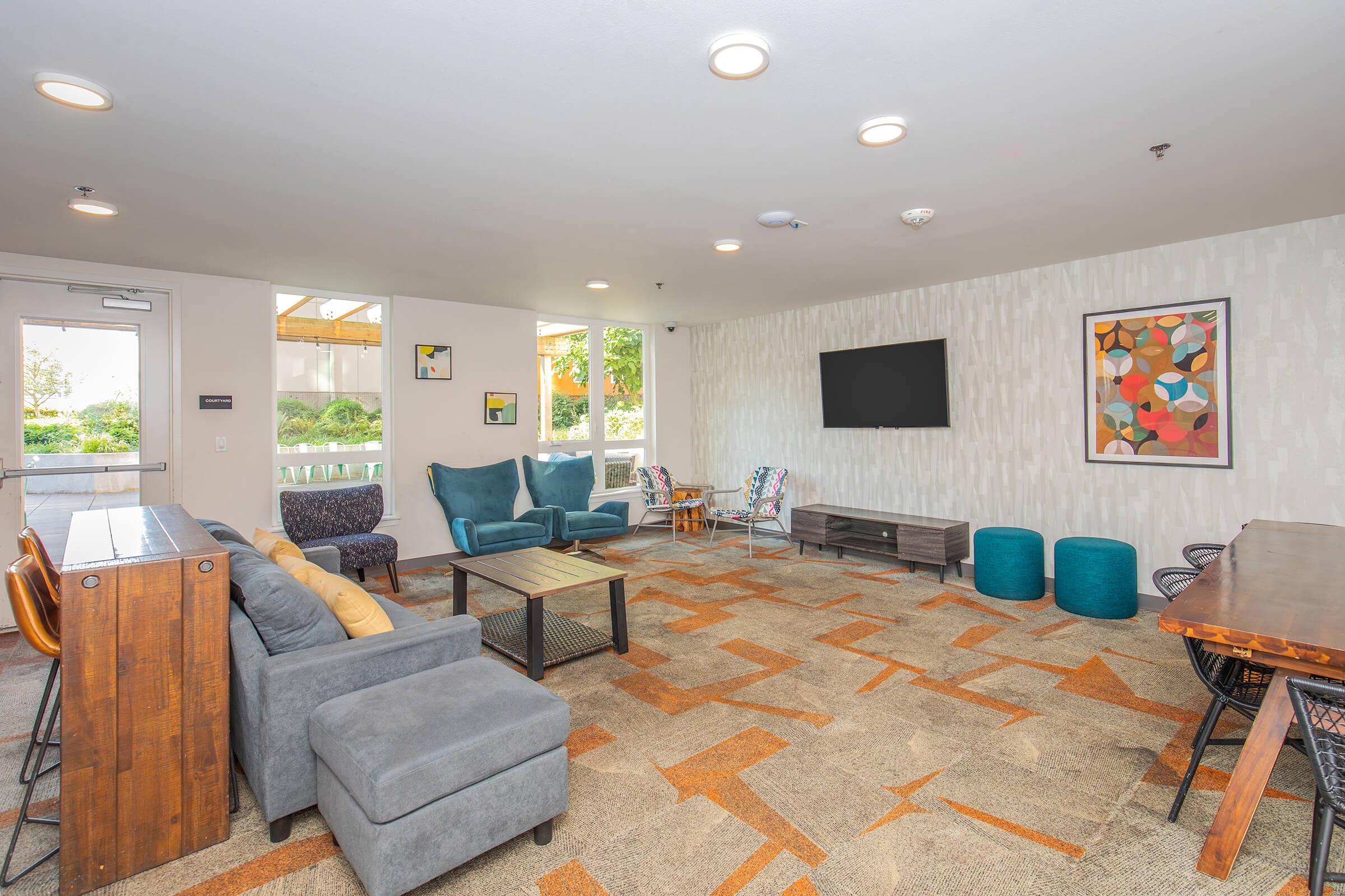 A modern, spacious lounge area with a mix of comfortable seating, including a gray sectional couch, colorful accent chairs, and a wooden coffee table. Large windows provide natural light, and a television is mounted on the wall. The floor features an abstract carpet design, enhancing the inviting atmosphere.