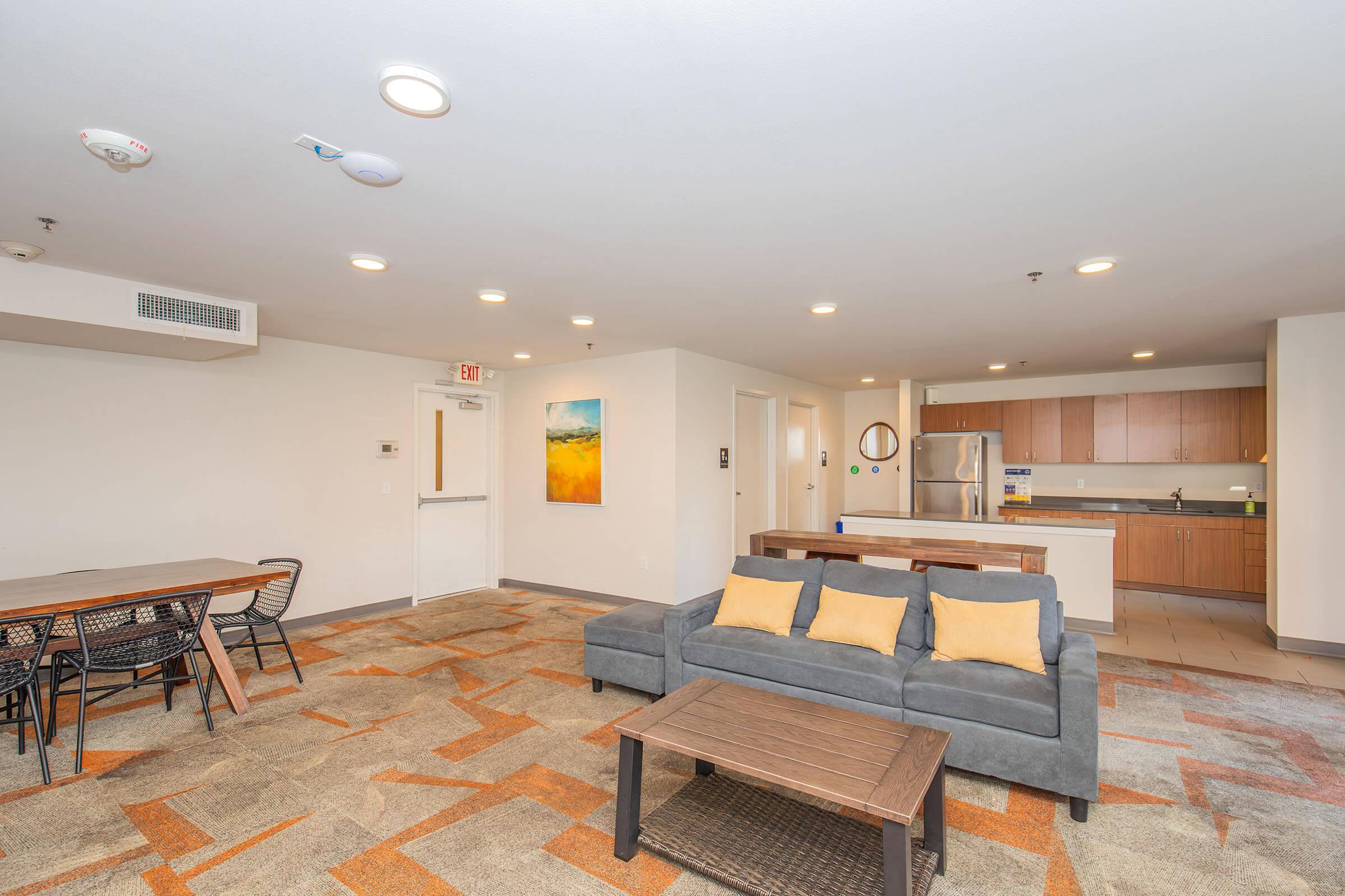 A modern living space featuring a cozy gray sofa with yellow cushions, a wooden coffee table, and a dining area with a rectangular table and chairs. The room has warm lighting, a kitchenette with appliances, and contemporary decor with an abstract painting on the wall.