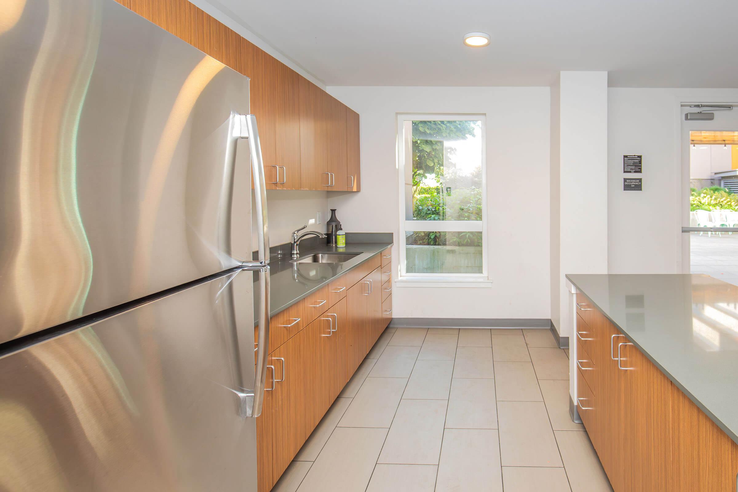 Modern kitchen featuring stainless steel appliances, wooden cabinetry, and a light-colored countertop. Natural light enters through a large window, highlighting the open space and tiled floor. The design is functional and contemporary, creating a welcoming atmosphere.